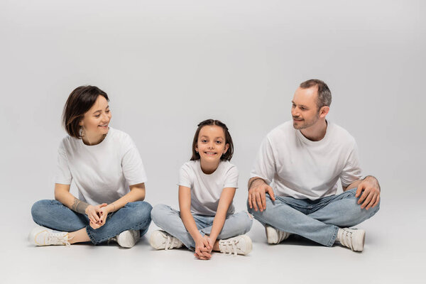joyous father and tattooed mother with short hair looking at cheerful preteen daughter while sitting with crossed legs in white t-shirts and blue denim jeans on grey background, Happy children's day 