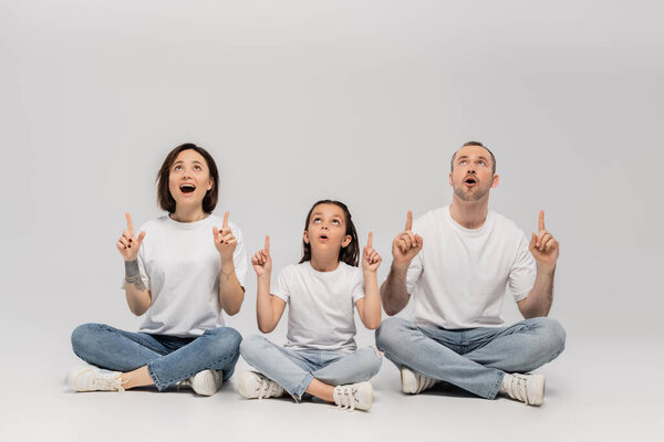 amazed family sitting with crossed legs in white t-shirts and blue denim jeans while pointing with fingers and looking up on grey background, father, tattooed mother and preteen daughter 