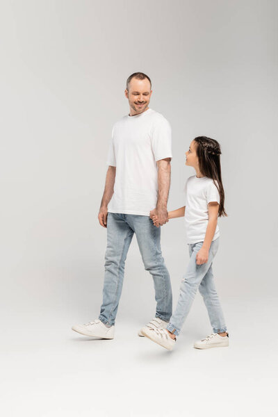 full length of cheerful father with bristle and preteen daughter in white t-shirts and blue denim jeans holding hands and walking together on grey background, Happy Father`s Day