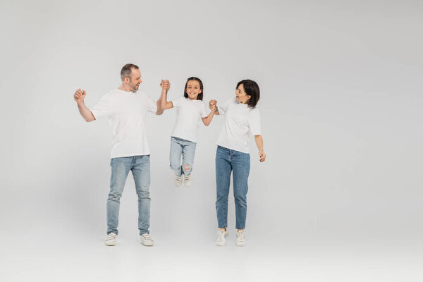 full length of happy parents in white t-shirts and blue denim jeans holding hands and lifting preteen daughter while standing together on grey background, International child protection day in June 