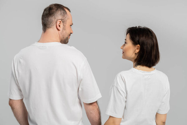 back view of cheerful and tattooed woman with short brunette hair looking at husband with bristle while standing together in white t-shirts isolated on grey background, happy couple 