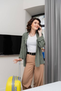 Esmer, sıradan kıyafetler, gömlek ve pantolon giyen, sarı valiz, pencere, gri perdeler ve otel odasındaki boş ekranlı lcd TV 'nin yanında dururken cep telefonuyla konuşan pozitif kadın.