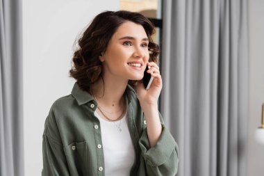 phone call, cheerful young woman with brunette hair wearing stylish shirt and smiling during conversation on smartphone in hotel room, travel lifestyle, cozy atmosphere clipart