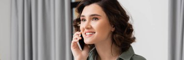 phone call, young and excited woman with brunette hair talking on mobile phone near grey curtains in contemporary and cozy hotel suite, leisure and travel, banner clipart