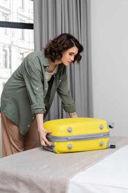 Genç ve çekici bir kadın, dalgalı esmer saçlı, dövmeli, şık gömlekli ve pantolonlu, sarı valizini açan, modern otel odasındaki gri perdeli, yatak ve pencereli.
