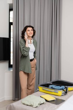 pozitif ve dövmeli kulaklıklı bir kadın cep telefonuyla konuşuyor gri perdelerin yanında, çantanın içinde seyahat gereksinimleri, not defterleri ve dizüstü bilgisayar var.