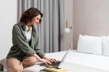 Dijital göçebelik, kablosuz kulaklıklı, gülümseyen bir kadın yatağında oturuyor ve dizüstü bilgisayarının yanında çalışıyor. Kalem akıllı telefon, beyaz yastık, duvar lambası ve otel odasındaki gri perdeler.