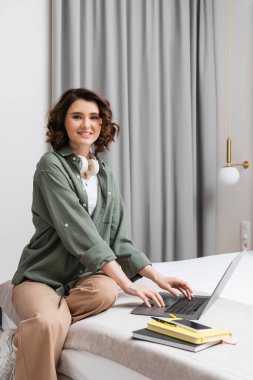 Kablosuz kulaklıklı neşeli dövmeli kadın yatakta oturuyor ve gri perdelerin yanındaki kameraya bakıyor, laptop, defterler, kalem ve akıllı telefon ile otelde boş ekran, dijital göçebelik 