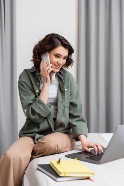 Kablosuz kulaklıklı neşeli bir kadın cep telefonuyla konuşuyor ve laptopla çalışıyor. Gri perdelerin, defterlerin ve kalemin yanında, rahat otel odasında, yatağın üzerinde oturuyor. 