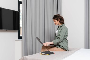 Dalgalı esmer saçlı, rahat ve modern otel süitinde, LCD TV 'nin yanında dizüstü bilgisayara, gri perdelere ve defterlere bakan neşeli bir kadın.