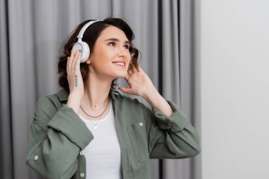 Modern otel odasındaki gri perdelerin yanında kablosuz kulaklıkla müzik dinlerken, dövmeli ve dalgalı esmer saçlı neşeli genç bir kadın.