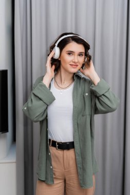 Dalgalı esmer saçlı ve dövmeli bir kadın kameraya bakarken kablosuz kulaklıkla modern otel odasındaki gri perdelerin yanında podcast dinliyor.