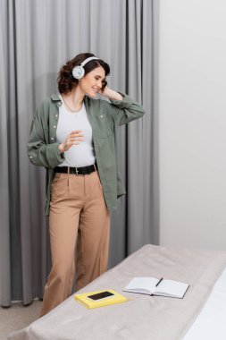 Kablosuz kulaklıkla müzik dinleyen, dalgalı esmer saçlı kaygısız dövmeli kadın. Gri perdelerin yanında duran akıllı telefon, boş ekran ve rahat otel odasındaki yatağın üzerinde defterler.