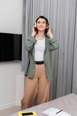 cheerful and tattooed woman in casual clothes adjusting wireless headphones and looking away while listening podcast near grey curtains, lcd tv, notebooks, pen and smartphone with blank screen in hotel room clipart