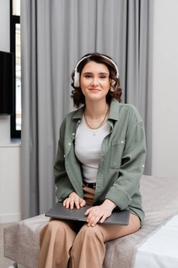 Dalgalı esmer saçlı mutlu bir kadın, gündelik kıyafetleriyle dizüstü bilgisayarla yatakta oturuyor, otel odasındaki online eğitim sırasında gri perdelerin yanında kablosuz kulaklıkla podcast dinliyor.