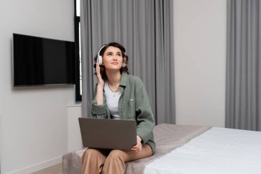 Kablosuz kulaklıkları ayarlayan ve otel odası, çalışma ve seyahat dersleri sırasında lcd TV ve gri perdelerin yanında dizüstü bilgisayarla yatağında oturan genç bir kadın.