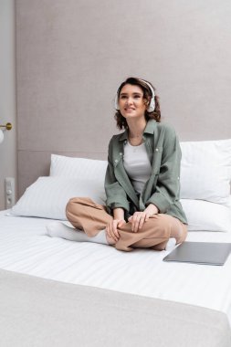 Kumral saçlı, günlük kıyafetleri ve kulaklıkları olan neşeli bir kadın bacak bacak bacak üstüne atmış bir şekilde yatakta oturuyor ve beyaz yastıkların yanında kablosuz kulaklıkla müzik podcast 'i dinliyor ve otel odasında dizüstü bilgisayar kullanıyor.