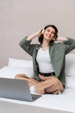 Dalgalı esmer saçlı, gözleri kapalı, kablosuz kulaklıkla müzik yayını dinleyen kaygısız bir kadın otel odasında dizüstü bilgisayar ve beyaz yastıkların yanında oturuyor.