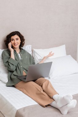 Dalgalı esmer saçlı ve dövmeli neşeli genç bir kadın, akıllı telefondan konuşurken otel odasındaki beyaz yastıkların yanında dizüstü bilgisayarla otururken el kol hareketi yapıyor.