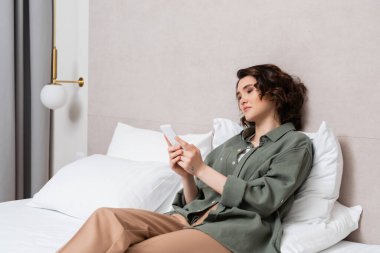 young woman with wavy brunette hair and tattoo sitting on bed in casual closed and browsing internet on mobile phone near white pillows and wall sconce in comfortable hotel room, leisure and travel clipart