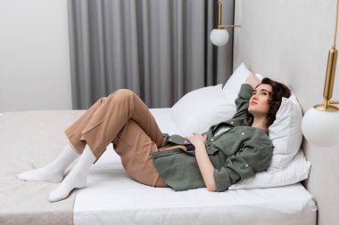 Dalgalı esmer saçlı, beyaz yastıkların yanında günlük giysiler içinde uzanan, rüya gibi ve kaygısız bir kadın. Modern otel odasının rahat atmosferinde, duvar lambaları ve gri perdeler.