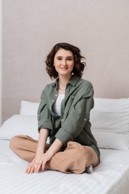 Dalgalı esmer saçlı kaygısız bir kadın gündelik giysiler içinde, bacak bacak üstüne atmış beyaz yastıkların yanındaki kameraya ve modern otel odasındaki gri duvara bakıyor.