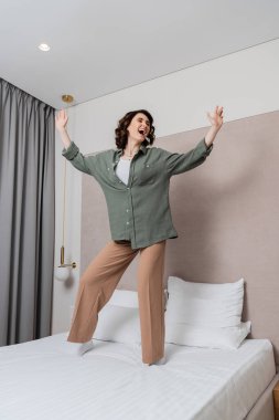 Uzun boylu ve neşeli bir kadın, gündelik giysiler içinde ve dalgalı esmer saçlarıyla beyaz yastıkların yanında dans ediyor ve şarkı söylüyor, gri perdeler ve modern otel odasındaki duvar lambaları.