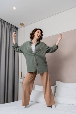 Dalgalı esmer saçlı genç bir kadın, günlük kıyafetler giyiyor, beyaz yastıkların yanında dans ediyor ve şarkı söylüyor, duvar lambası ve rahat otel odasındaki gri perdeler