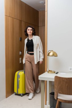 Modern otel odasında sarı bavul, altın masa lambası, sandalye ve çöp kovası ile ayakta duran şık, gündelik giysiler içinde mutlu bir kadın.