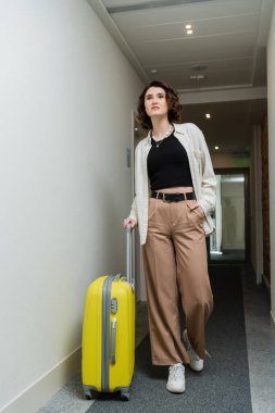 Kendine güvenen, esmer, dalgalı saçlı, beyaz tişörtlü, siyah bluzlu, cebinde bej pantolonlu, modern otelin koridorunda sarı valizli bir kadın.