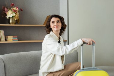 Dalgalı esmer saçlı, dövmeli, şık kıyafetleri olan, sarı seyahat çantasının yanındaki kanepede oturan, modern otelin lobisinde raflarında çiçekler olan genç ve mutlu bir kadın.