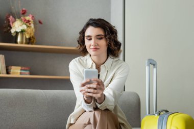 Kumral saçlı genç bir kadın seyahat çantasının yanındaki koltukta otururken akıllı telefon kullanıyor. Kitaplarla dolu raflar ve otel lobisindeki bulanık arka planda çiçekli vazolar, sosyal ağlar. 