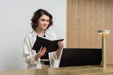 positive receptionist in trendy casual clothes, with wavy brunette hair standing with notebook near computer monitor and lamp on front desk in lobby of contemporary hotel  clipart