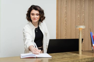 Modern otel lobisinde resepsiyon masasında çalışırken bilgisayar monitörü ve lambanın yanında not defterine dalgalı esmer saçlarıyla şık günlük giysiler içinde arkadaş canlısı kapıcı.