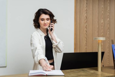 joyful receptionist with wavy brunette hair working at front desk, writing in notebook and talking on telephone near computer monitor in lobby of modern hotel, hospitality industry clipart
