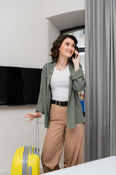 positive woman with brunette hair, in casual clothes, shirt and pants, talking on mobile phone while standing near yellow suitcase, window, grey curtains and lcd tv with blank screen in hotel room