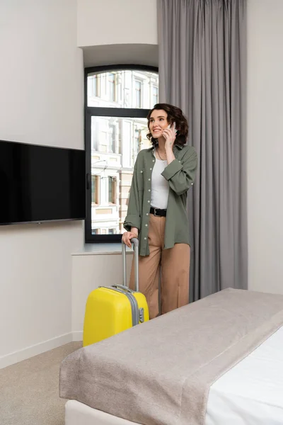 stock image business travel, happy and charming woman with wavy brunette hair, in casual clothes talking on smartphone near window with grey curtains, bed, yellow suitcase and lcd tv in hotel room