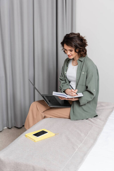 carefree young woman writing in notebook and looking at laptop while sitting on bed near grey curtain, notepad and smartphone with blank screen in hotel suite, freelance lifestyle, work and travel