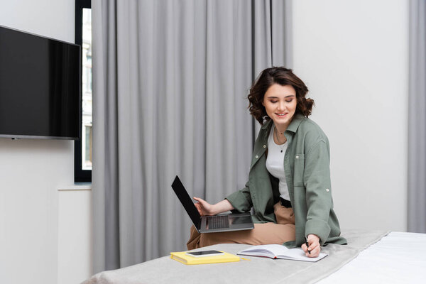 remote work, smiling woman with wavy hair writing in notebook and looking at camera while sitting near grey curtains, notepad and smartphone with blank screen on bed in modern hotel room