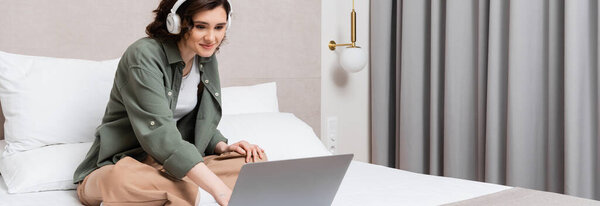 довольная молодая женщина с волнистыми брюнетками, в беспроводных наушниках, смотрящая фильм на ноутбуке, сидя на кровати со скрещенными ногами возле серых занавесок и настенного булочки в гостиничном номере, баннер