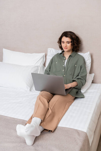 full length of young woman in casual clothes, with wavy brunette hair working on laptop while sitting on bed near white pillows and grey wall in comfortable hotel room