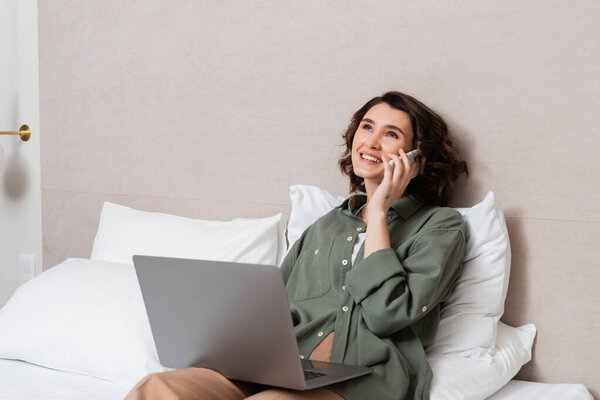 smiling woman with wavy brunette hair wearing casual clothes, holding laptop and talking on mobile phone while sitting on bed near white pillows in cozy hotel room, work-life integration