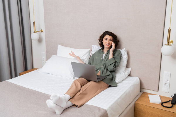 full length of joyful tattooed woman in casual clothes sitting on bed with laptop near white pillows, wall sconces and grey curtains, gesturing during conversation on mobile phone in modern hotel room