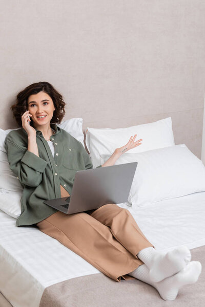 cheerful young woman with wavy brunette hair and tattoo, in casual clothes gesturing during conversation on smartphone while sitting on bed with laptop near white pillows in hotel room