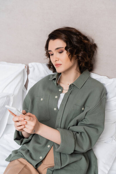 young woman with wavy brunette hair and tattoo browsing internet on mobile phone while sitting on bed near grey wall and white pillows in cozy atmosphere of hotel room, leisure and travel