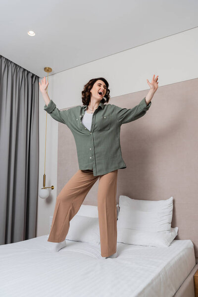 full length of young and overjoyed woman in casual clothes and with wavy brunette hair dancing and singing on bed near white pillows, grey curtains and wall sconce in modern hotel suite
