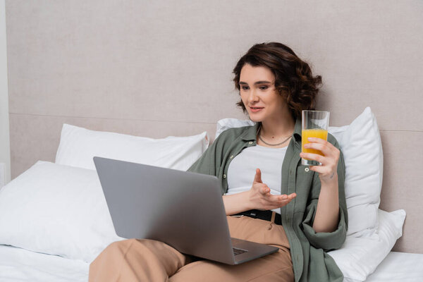 smiling woman with wavy brunette hair holding glass of fresh orange juice and gesturing during video call on laptop while sitting on bed near white pillows and grey wall in hotel room