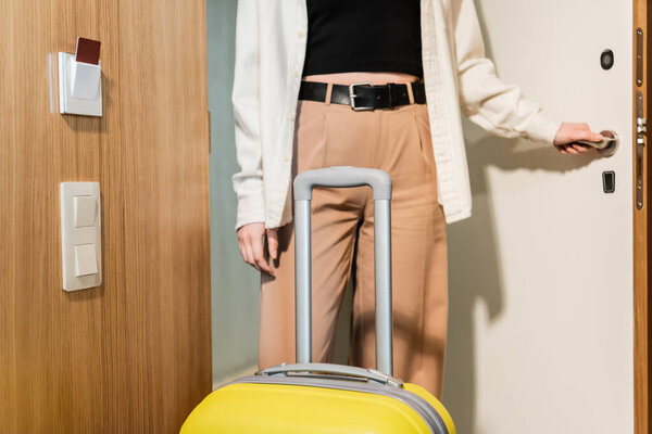 cropped view of young and stylish woman in white shirt and beige pants opening entrance door in modern hotel near yellow travel bag and keycard reader, secure entry, travel lifestyle, check-in 