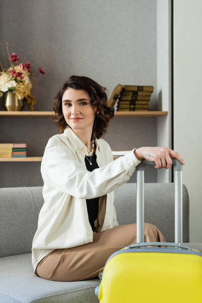 carefree tattooed woman with wavy brunette hair, in stylish casual clothes looking at camera while sitting on couch with yellow suitcase, near shelves with books and vase with flowers in hotel lobby
