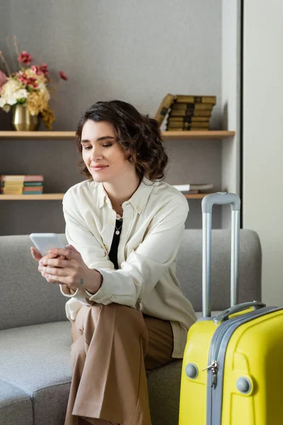 喜欢纹身的女人 一头卷曲的黑发 坐在沙发上 靠近黄色旅行袋 在智能手机上边看书边浏览互联网 在酒店大堂背景模糊的花瓶上挂着花 — 图库照片